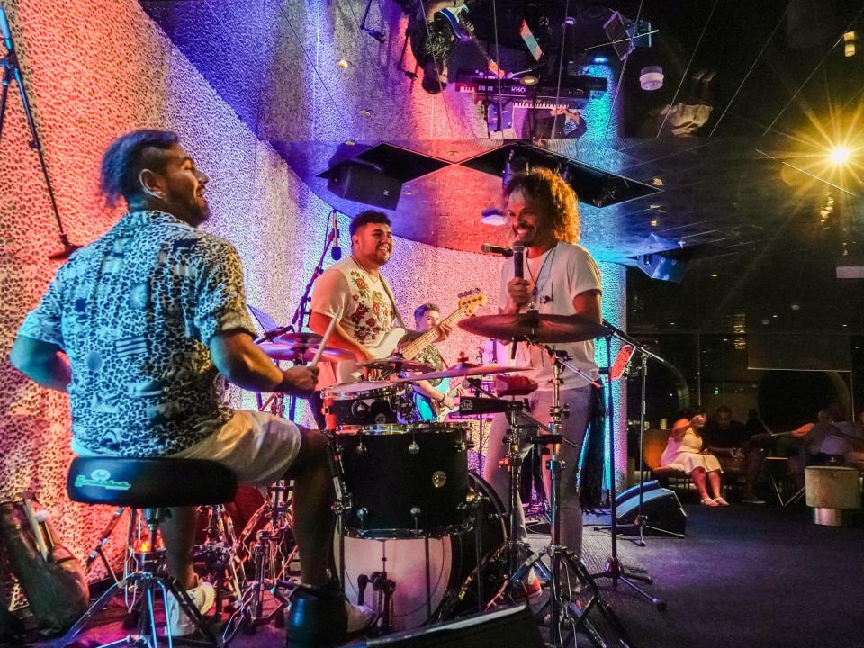 A dark room with a band playing in front of colorful lighting on the left and people seating watching on the right