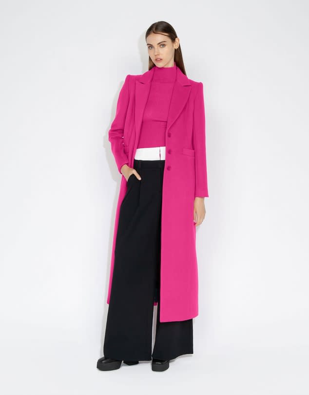 A model with long dark hair wears an Ultra Pink Maxi Coat and pink turtleneck and wide black pants and chunky boots.