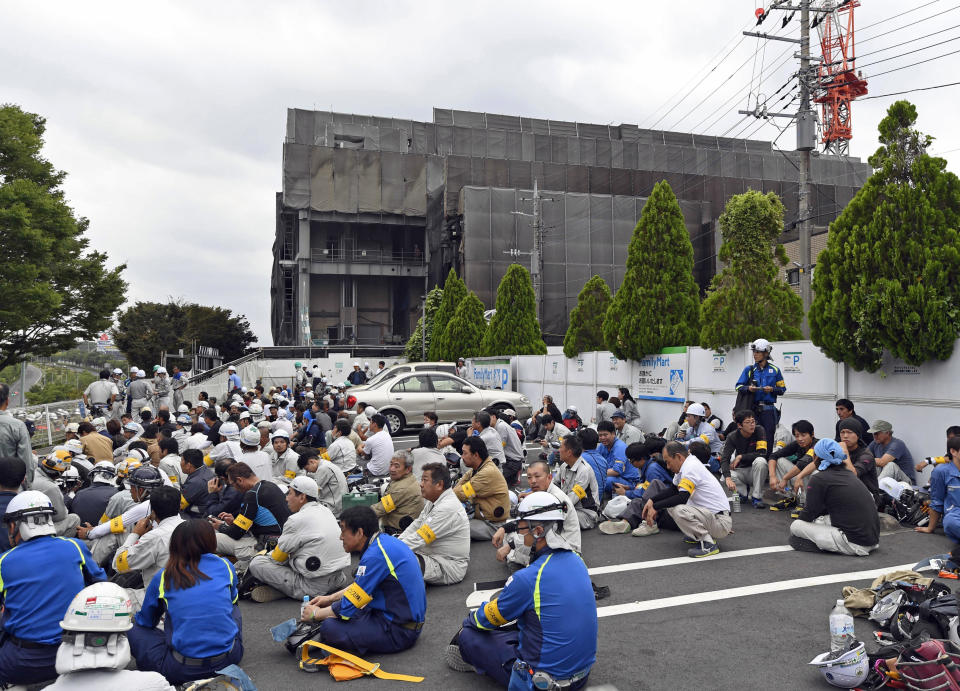 Workers who fled from the site of a fire, seen in the background, take shelter in Tama city, Tokyo's western suburbs, Thursday, July 26, 2018. The fire broke out Thursday afternoon, injuring a number of people at the building being constructed. (Kyodo News via AP)