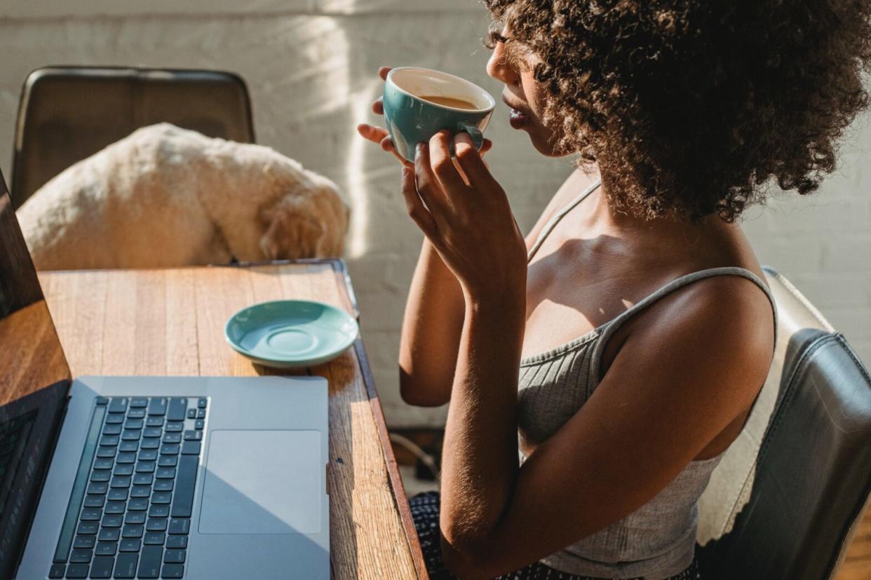 Black woman drinking coffee in front of computer with dog in chair