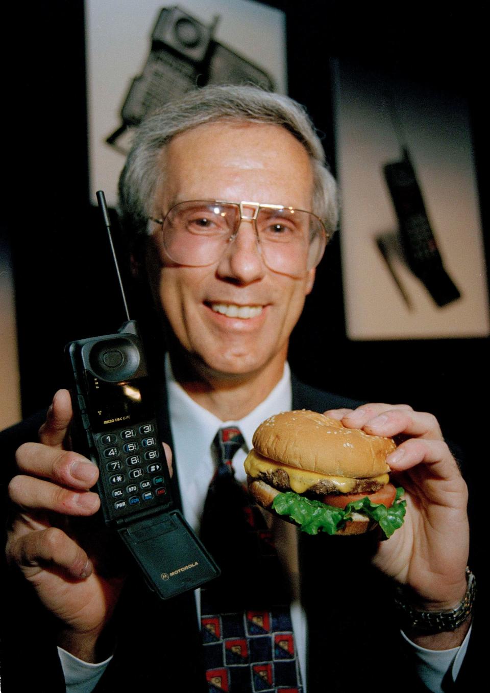man with gray hair wearing a suite holding a Motorola MicroTac Elite cell phone in one hand, and a burger in his other hand to compare the sizes