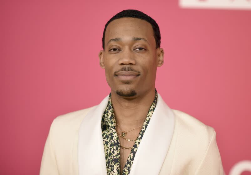 Tyler James Williams poses in a white blazer and patterned shirt against a pink backdrop.