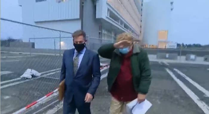 Two men wearing face masks walk in a parking lot, one covering his face with his hand