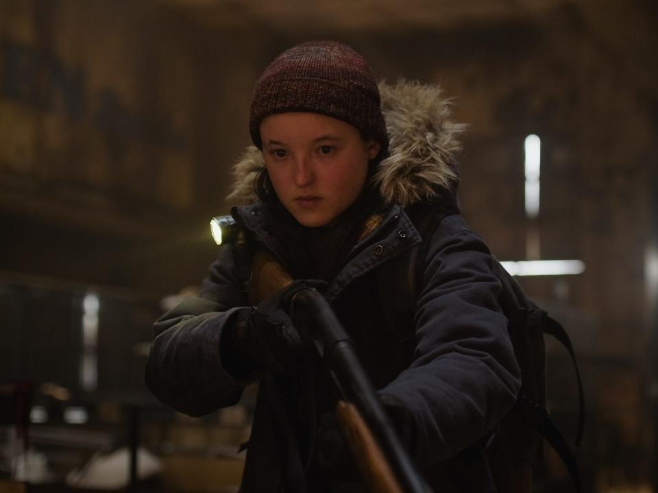 bella ramsey as ellie in the last of us season two, wearing a winter coat, a hat, and intently holding a gun