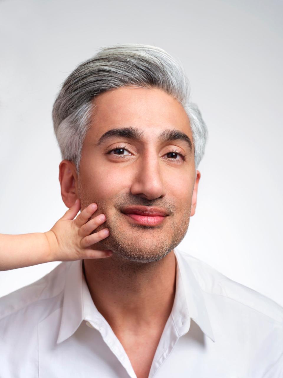 Tan France smiles in a white shirt as a baby's hand rests on his cheek.