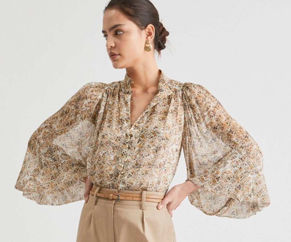 A woman wears a beige and sage patterned sheer shirt with billowy sleeves against a white backdrop.