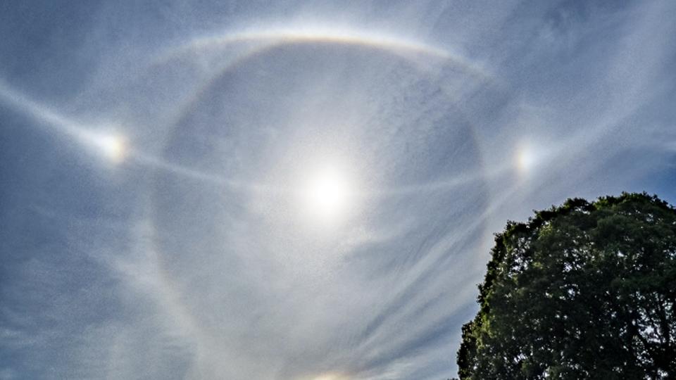 A blue sky with the sun surrounded by a white circle bisected with a white line and two bright spots either side of the circle.