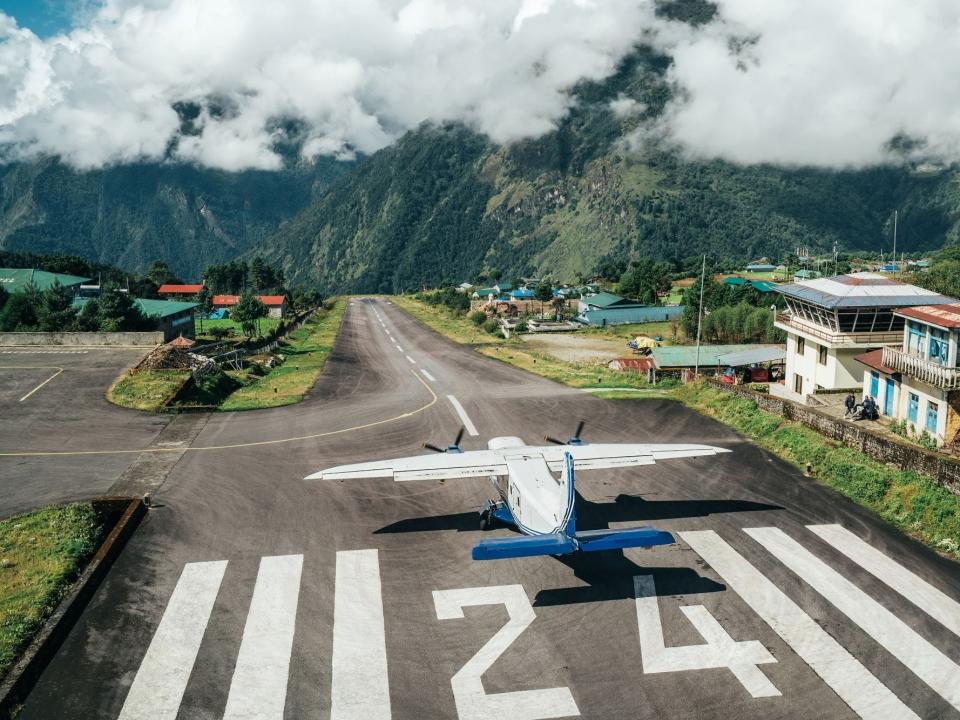 A plane getting to takeoff from runway 24 at Tenzing-Hillary Airport in Nepal with clouds, mountains, and greenery in the background.