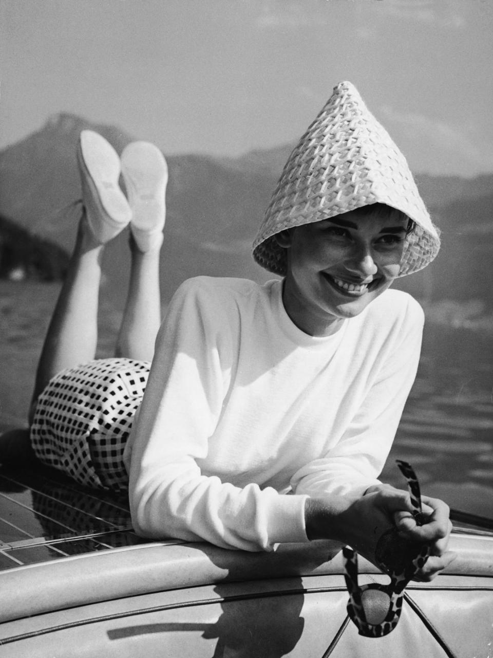 <p>Audrey Hepburn lays across the bow of a motorboat during a day on the water. The stylish actress wears a gingham print skirt, white mock turtleneck, and woven sun hat. </p>