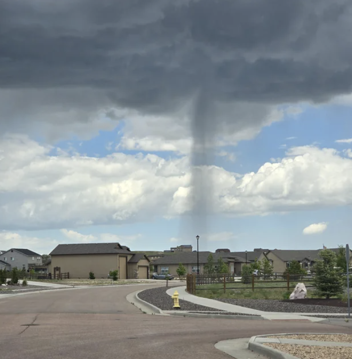 A thin tornado descends from dark clouds over a suburban neighborhood, with houses, a street, and a fire hydrant visible in the foreground