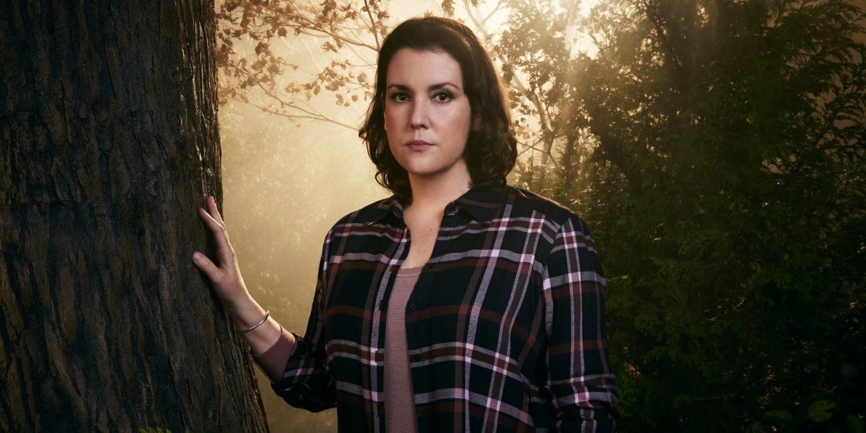 yellowjackets star melanie lynskey, in character as shauna, stands in a forest with her right hand resting on a tree trunk