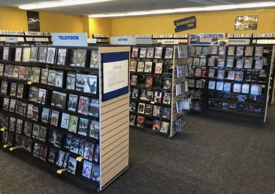 Rows of DVD shelves in a Blockbuster store, with sections labeled "Television," "Action," "New Releases," and "International."