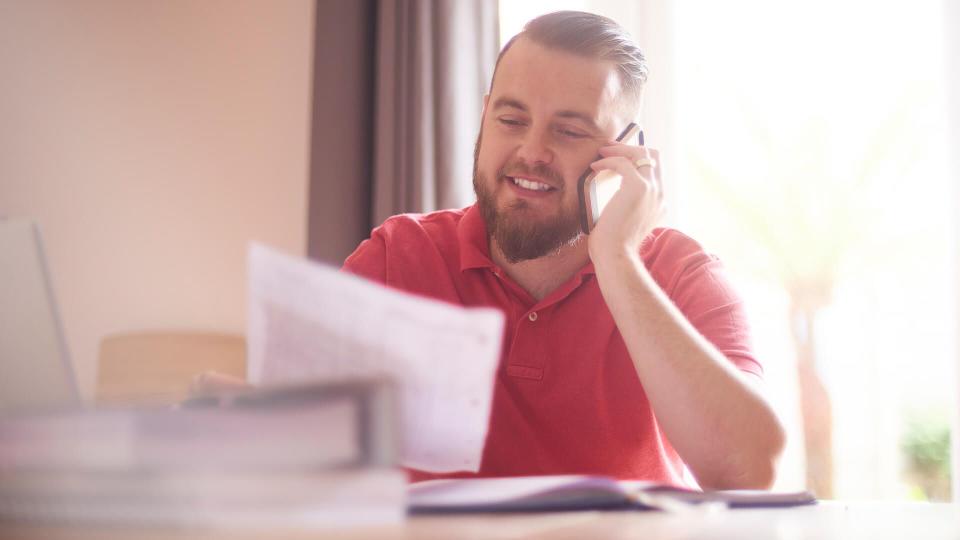 A young male possibly a single parent or small business owner sits at home talking to the bank or finance company on the phone.