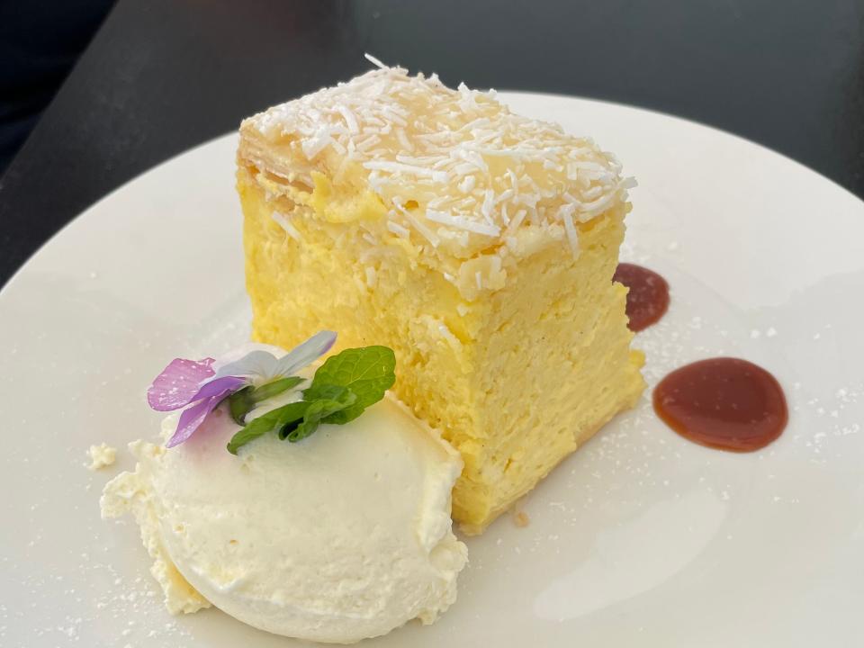 A white plate with a yellow custard square covered with white sprinkles next to a scoop of vanilla ice cream with a flower and herbs on top.