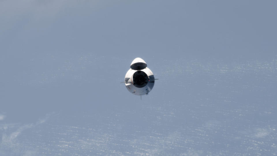 a white space capsule approaches the international space station, with a few clouds over the ocean in the background