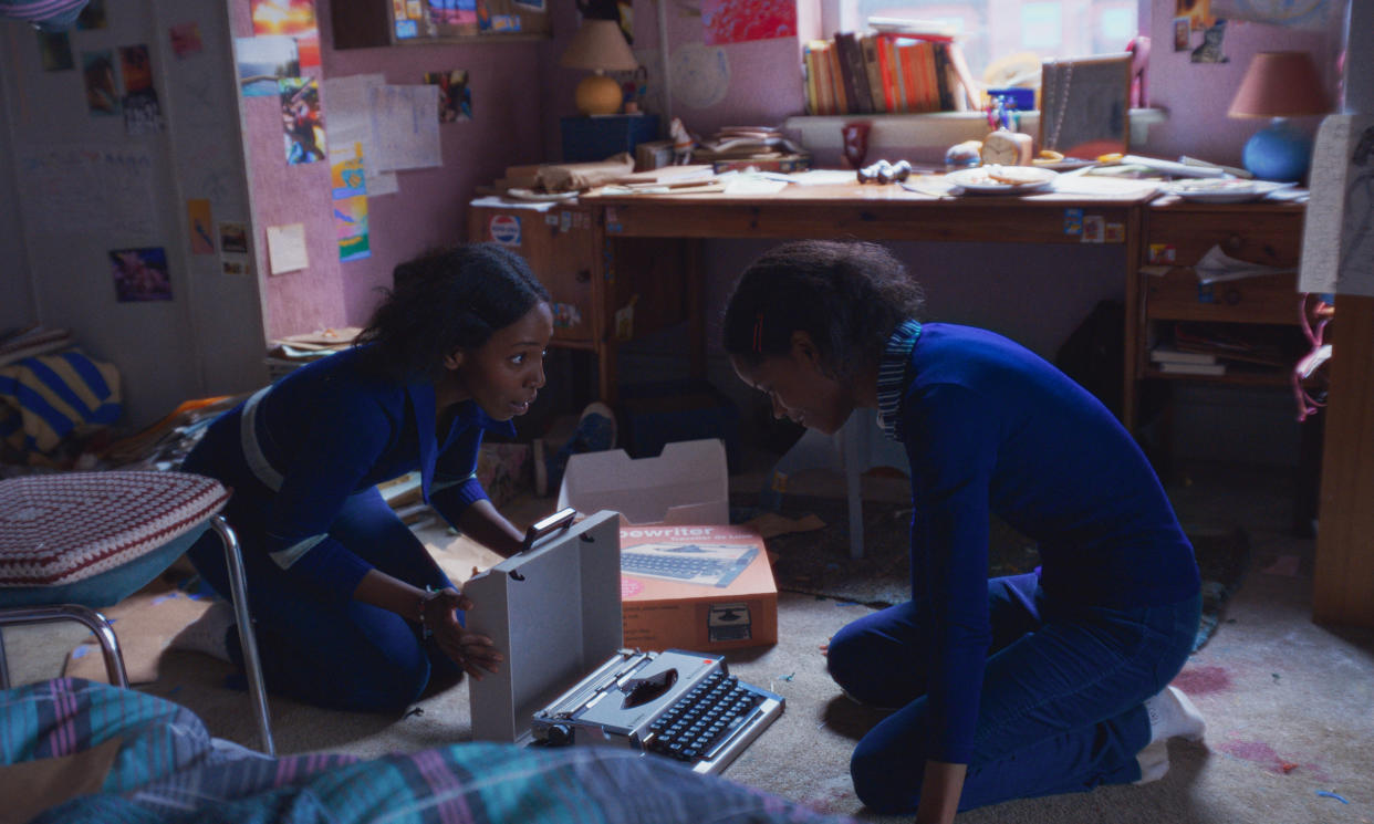 Tamara Lawrance as Jennifer Gibbons and Letitia Wright as June Gibbons open a new typewriter from its box in their bedroom.