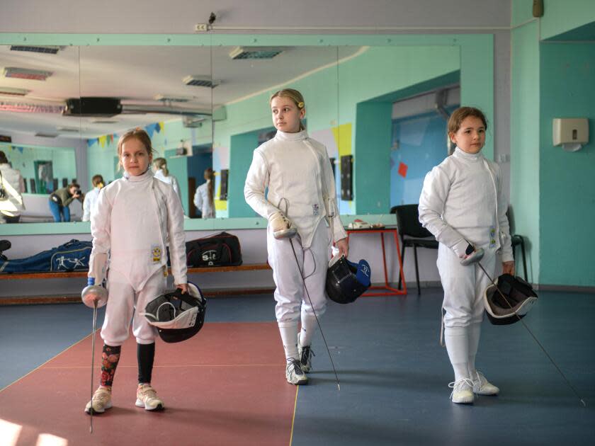 Three girls stand in fencing uniforms, holding helmets and equipment.