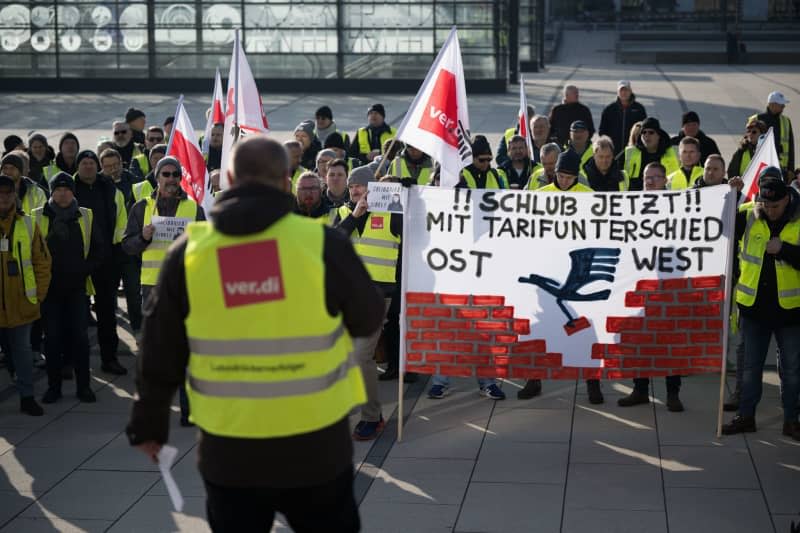 Strikers stand with a banner reading "No more tariff differences East West" during a rally at Berlin Brandenburg Airport. With renewed warning strikes by several professional groups, the Verdi trade union is paralyzing important parts of German air traffic on Thursday and Friday. Sebastian Gollnow/dpa