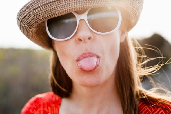 Mature woman wearing sunglasses and sunhat sticking out tongue