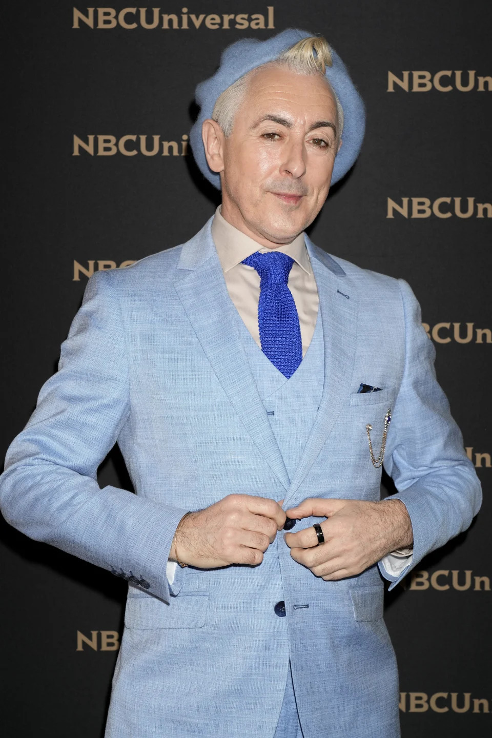 Alan Cumming poses in a light suit, tie, and hat at an NBCUniversal event