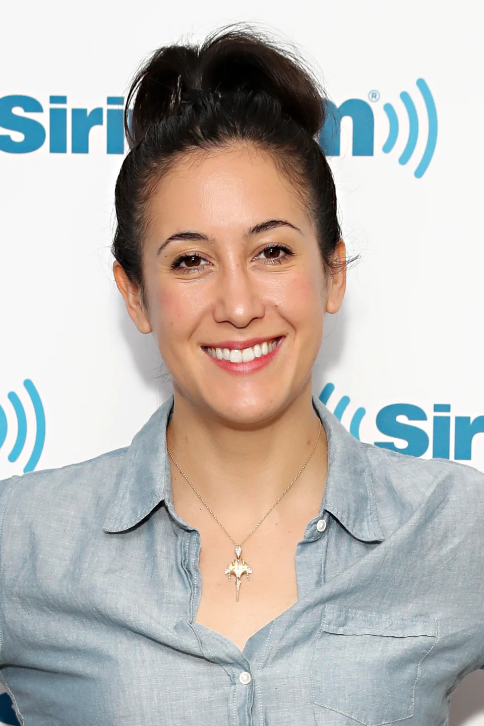 A person with dark hair in a bun, wearing a button-up shirt and a star-shaped necklace, smiling in front of a SiriusXM logo backdrop