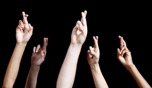 'Five mixed hands are raised against a black background, all with fingers hopefully crossed, averting bad luck.'