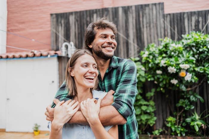 A happy couple, names unknown, standing outdoors. The man is hugging the woman from behind and they are both smiling. They are wearing casual clothes