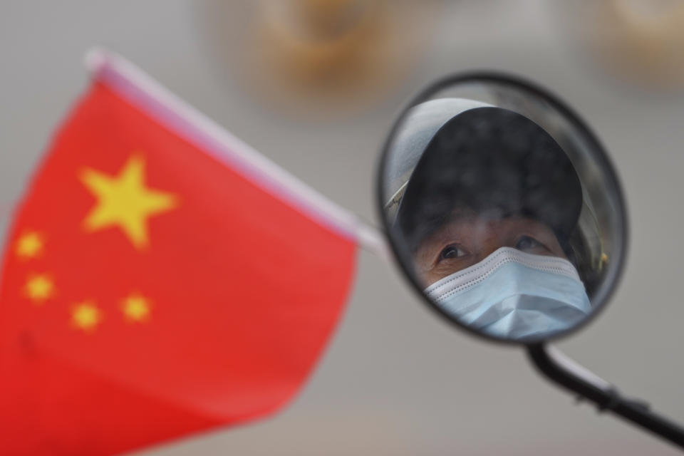 A woman wearing a face mask to help protect from the coronavirus is reflected in her wing mirror of her electric-powered scooter tied with a national flag as she waits at an intersection during the morning rush hour, Monday, April 11, 2022, in Beijing. (AP Photo/Andy Wong)