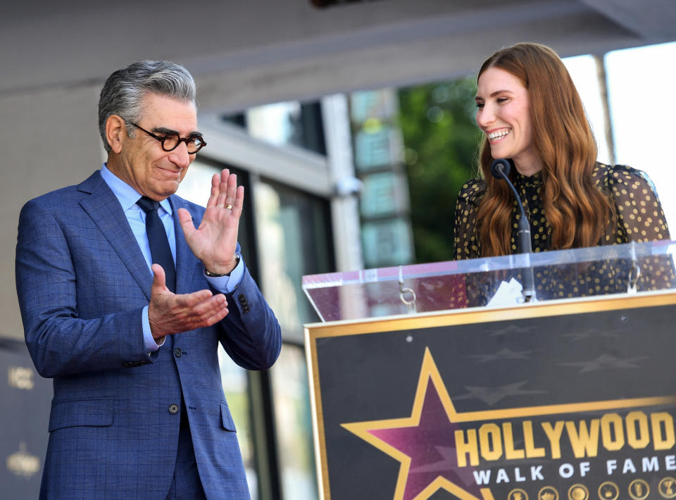 sarah speaking behind a podium while eugene stands and claps beside her