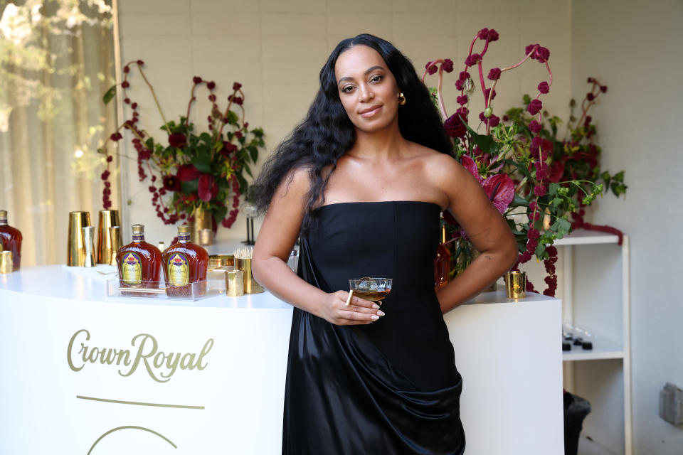 Solange Knowles is at an event, dressed in a strapless black dress, holding a drink. She stands in front of a display with Crown Royal bottles and flower arrangements