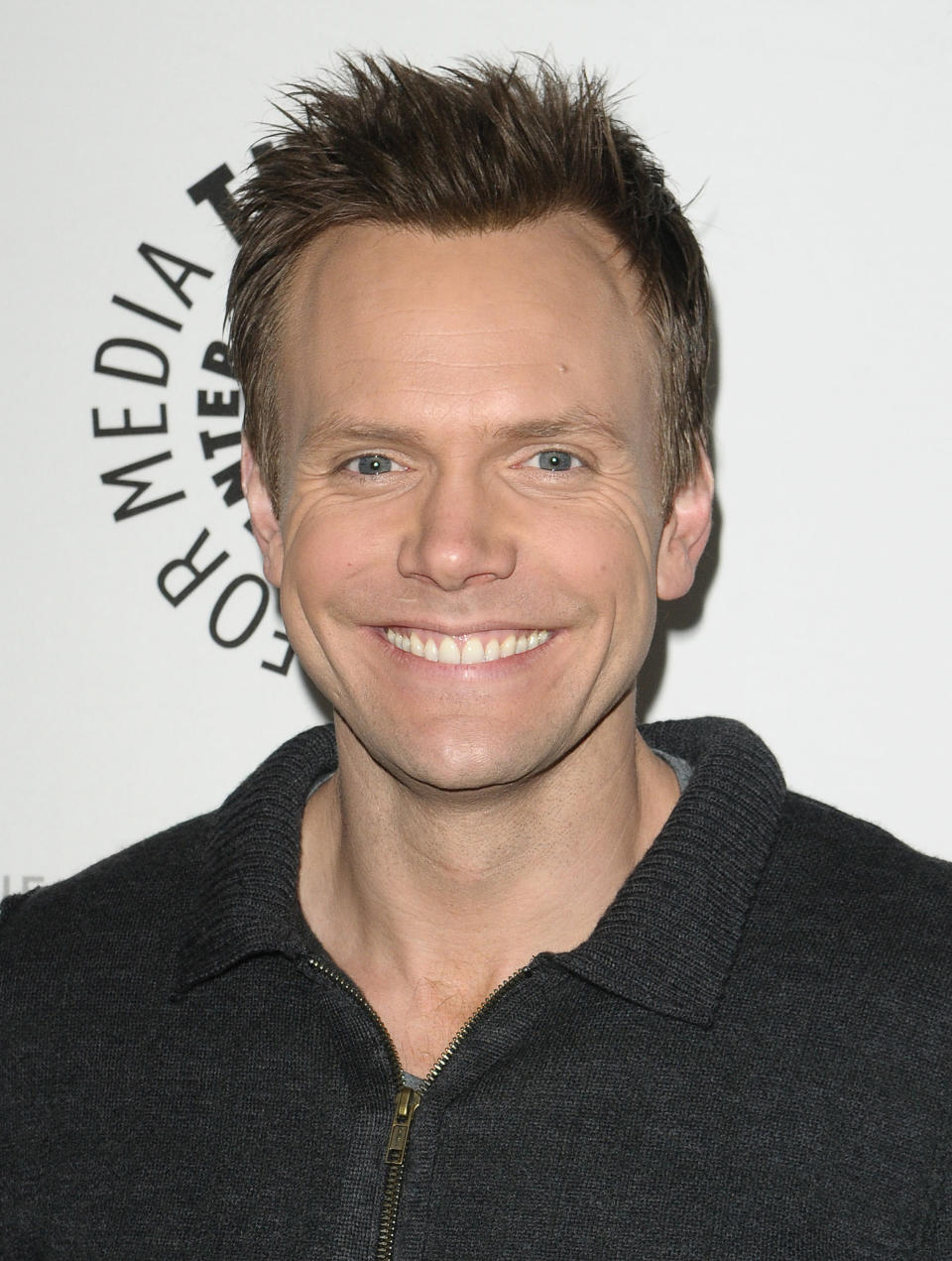 A closeup of Joel McHale smiling on the red carpet of a media event. He's wearing a zippered sweater