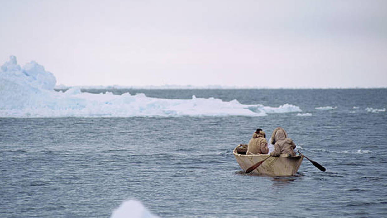  Two indigenous Arctic peoples paddle through icy waters in a traditional canoe-style boat. 