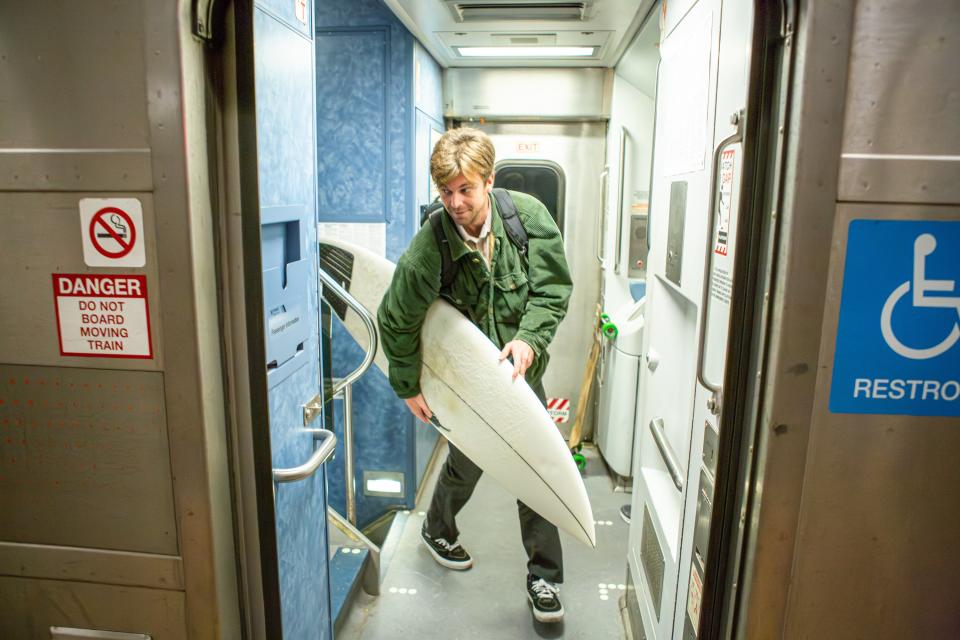 A thirtysomething white guy, the author, carries a surfboard through a train.