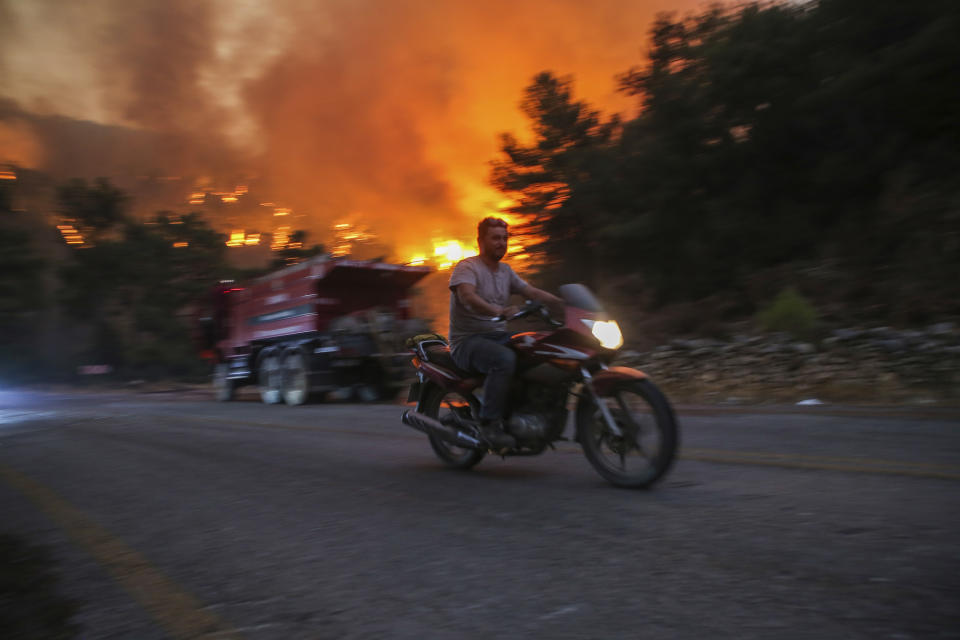 A man rides away from an advancing fire that rages Cokertme village, near Bodrum, Turkey, Monday, Aug. 2, 2021. For the sixth straight day, Turkish firefighters battled Monday to control the blazes that are tearing through forests near Turkey's beach destinations. Fed by strong winds and scorching temperatures, the fires that began Wednesday have left eight people dead. Residents and tourists have fled vacation resorts in flotillas of small boats or convoys of cars and trucks. (AP Photo/Emre Tazegul)