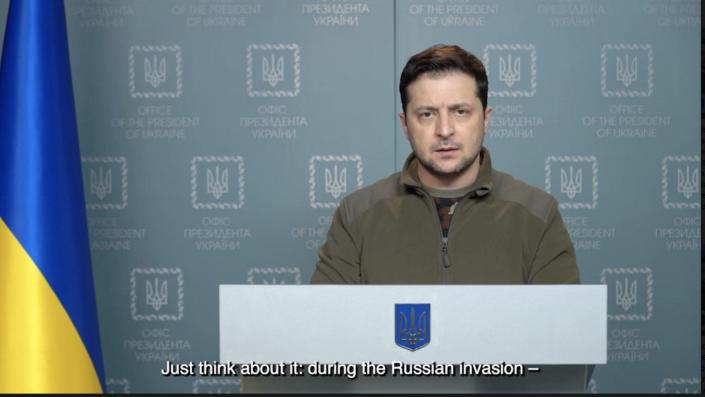 President Volodymyr Zelensky in a video address released February 28, 2022. He is at a podium with a Ukrainian flag nearby.