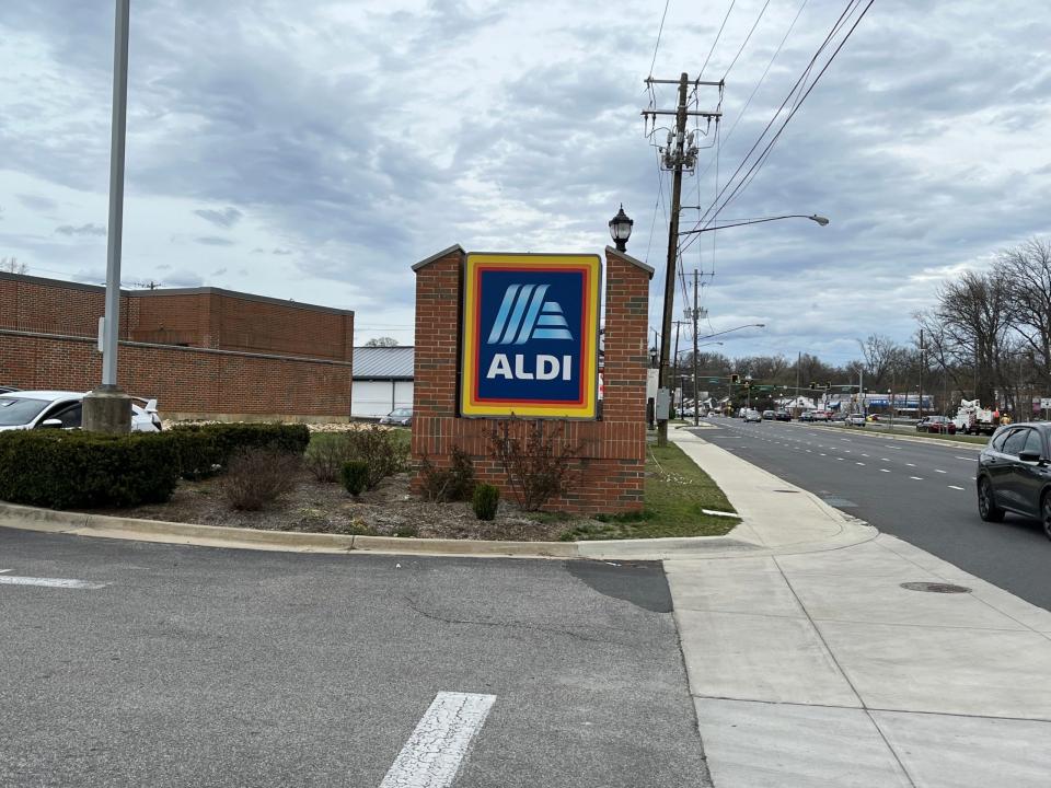 Ein blau-weißes Aldi-Schild mit einer roten, orangefarbenen und gelben Umrandung, die von Ziegelsteinen gehalten wird, ist neben einer viel befahrenen Straße zu sehen. Im Vordergrund ist die Einfahrt zum Aldi-Parkplatz zu sehen.