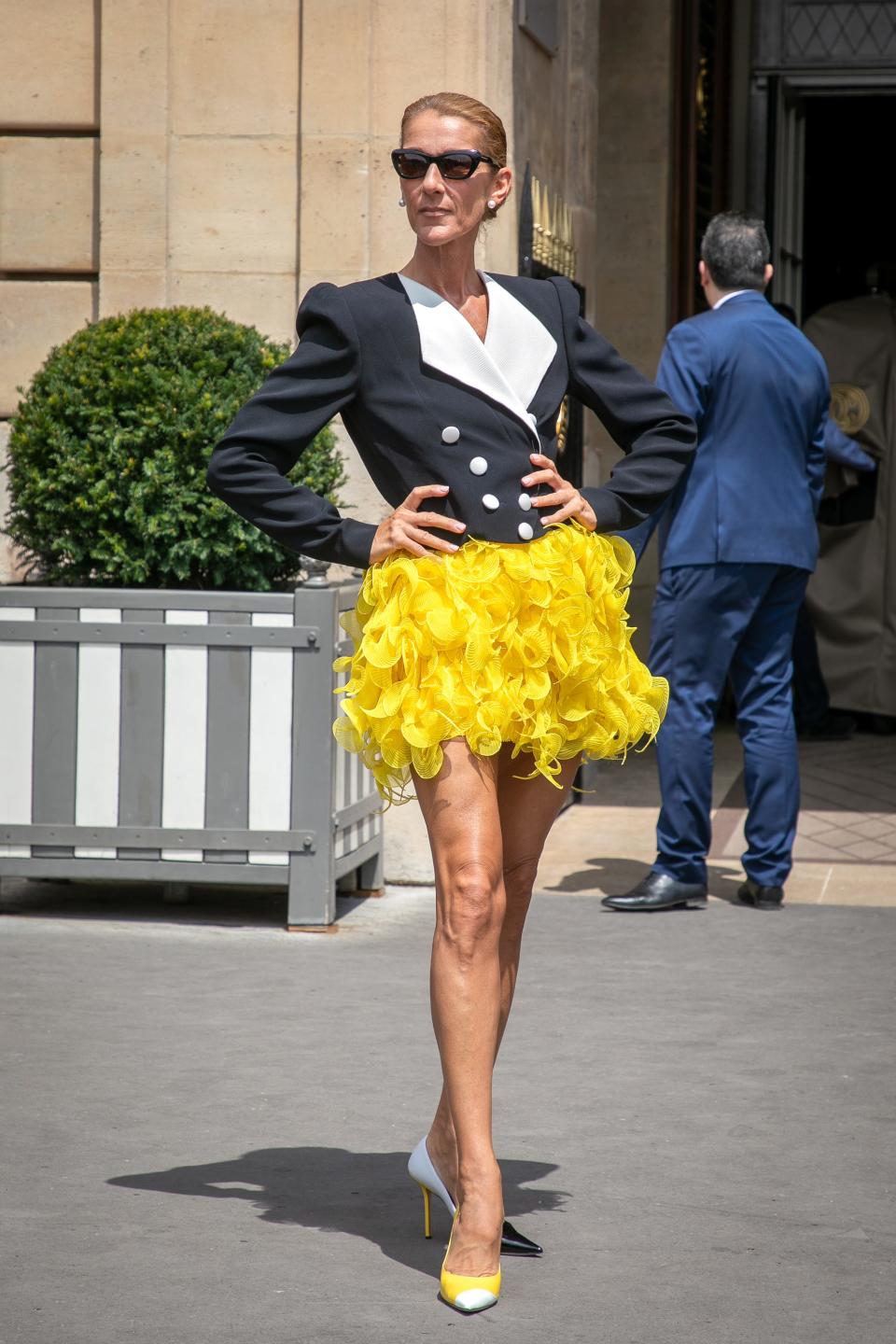 Celine in a black and white tuxedo-lke jacket, a feathery yellow miniskirt, sunglasses, and heels.