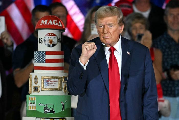 Donald Trump in a suit and red tie raises a fist next to a large decorated cake at an event