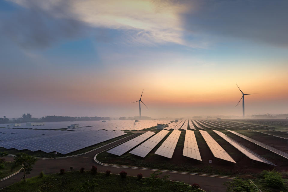 A field of solar panels with wind turbines in the background at dawn.