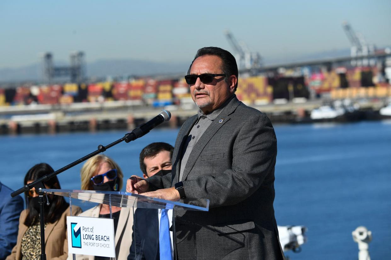 Ron Herrera at the microphone, with a body of water and an array of containers and cranes in the background.