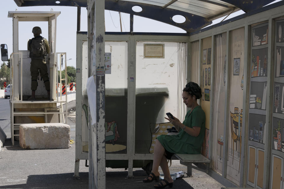 An Israeli soldier stands guard at a bus stop as a woman waits for a bus at the Gush Etzion junction, the transportation hub for a number of West Bank Jewish settlements, Thursday, June 9, 2022. Israeli settlers in the occupied West Bank may soon have a taste of the military rule that Palestinians have been living under for 55 years. A looming end-of-month deadline to extend legal protections to Jewish settlers has put Israel’s government on the brink of collapse and drawn widespread warnings that the territory could be plunged into chaos. (AP Photo/Maya Alleruzzo)