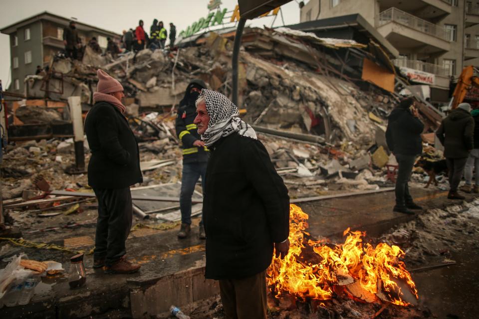 People warm themselves next to a collapsed building in Malatya, Turkey, on Feb. 7 2023, after a large earthquake shook the country. Aftershocks continued in the following weeks, causing some of the already damaged buildings to collapse.