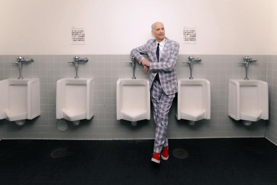 John Waters wears a plaid suit and stands amid bathroom urinals in a 2022 portrait.
