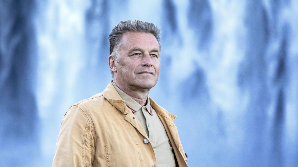 Chris Packham in a mustard jacket stands in front of a waterfall in Earth.