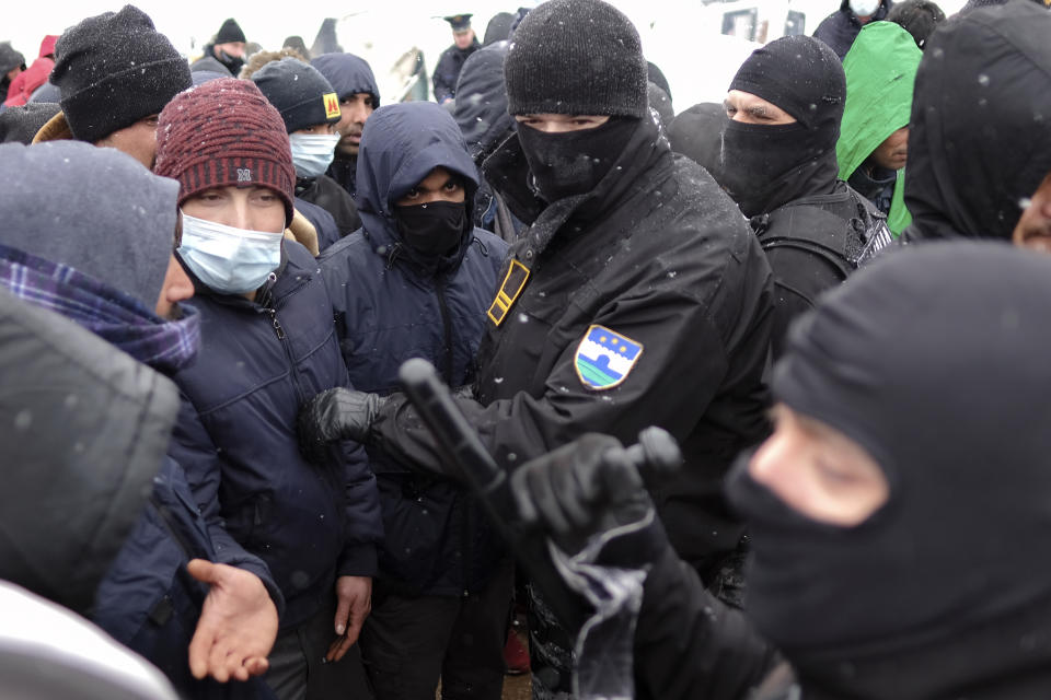 Bosnian police stand in front of migrants waiting in lines for food handouts during snowfall at the Lipa camp, outside Bihac, Bosnia, Friday, Jan. 8, 2021. A fresh spate of snowy and very cold winter weather on has brought more misery for hundreds of migrants who have been stuck for days in a burnt out camp in northwest Bosnia waiting for heating and other facilities. (AP Photo/Kemal Softic)