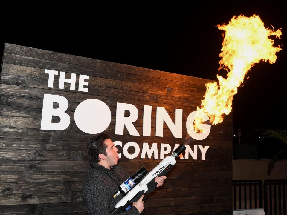 A man stands in front of a sign that says "The Boring Company" and brandishes a flamethrower, shooting flames into the air.