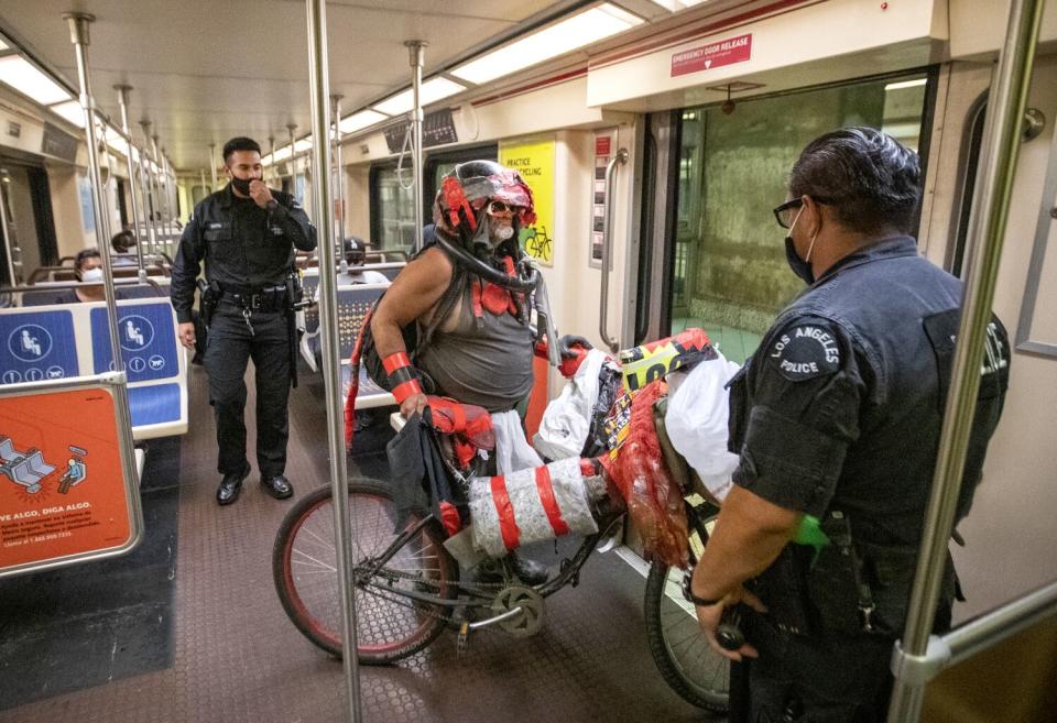 Two police officers stand on each side of a person in a helmet and with a bicycle loaded with various items.