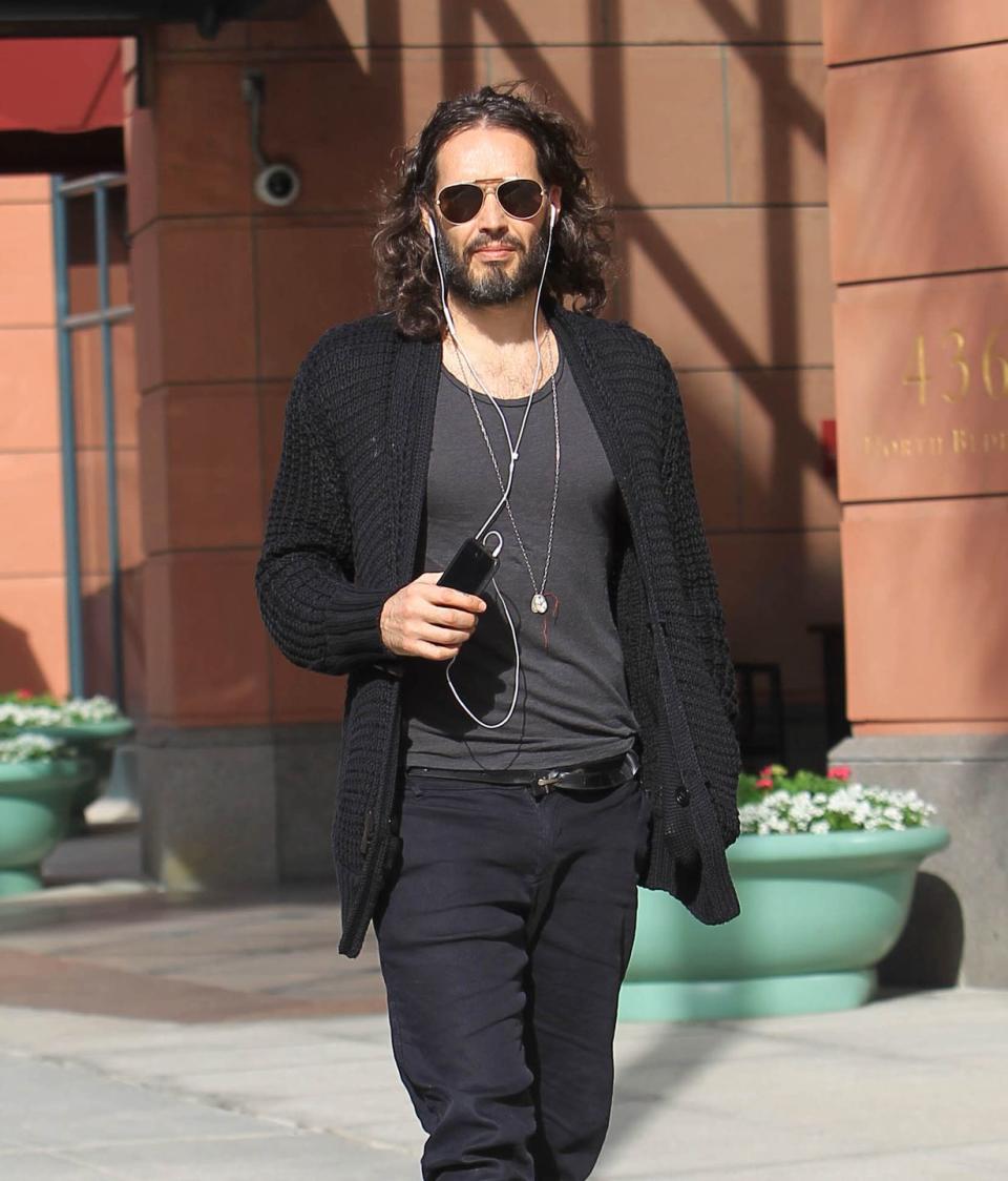 russell brand pictured in 2018 walking down the street, he is wearing sunglasses and a black and grey outfit, and has earphones in his ears