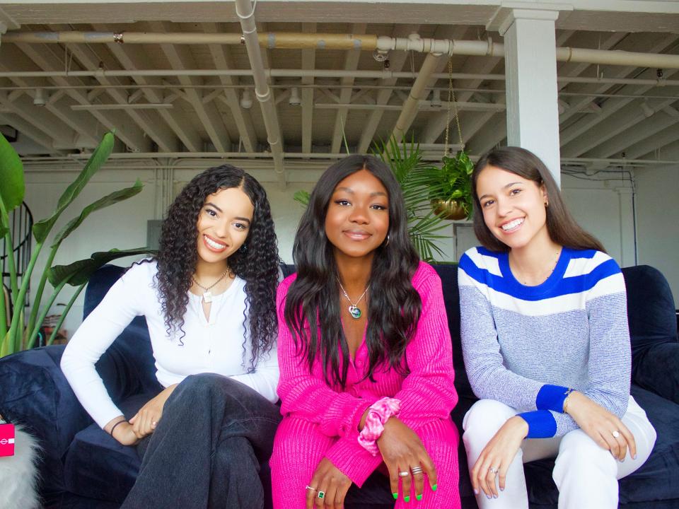 Three girls sit, one wearing a white shirt, the other in all pink and the third in a gray, white and blue sweater