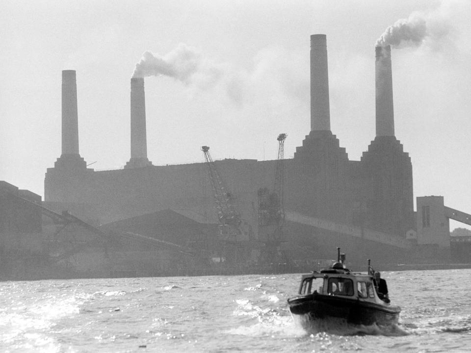 A black-and-white photo of a small boat on the river Thames with Battersea Power Station in the background, a rectangular building with four tall chimneys at its corners. Two of the chimneys are belching smoke.